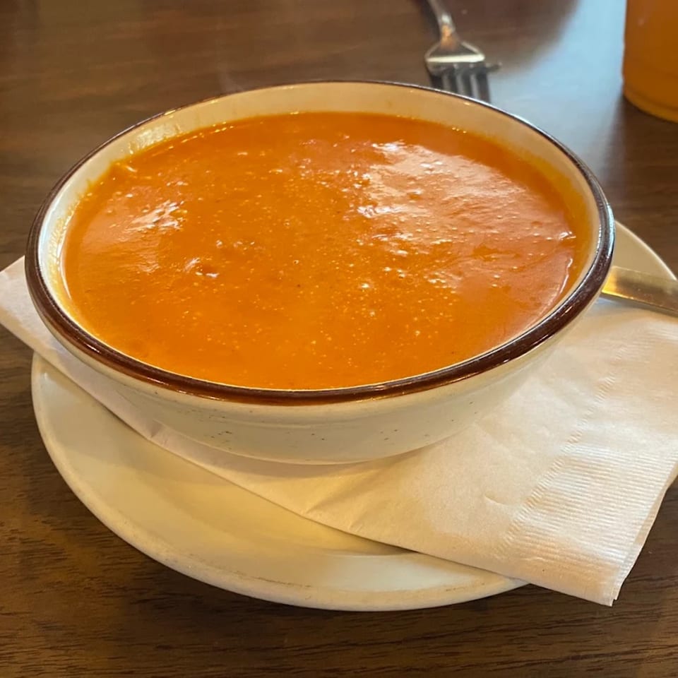 A bowl of vibrant orange Indian tomato soup, served on a white plate with a napkin.