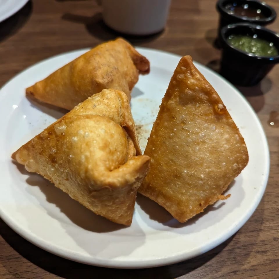 Three golden-brown Indian samosas on a white plate with two small bowls of dipping sauces in the background.