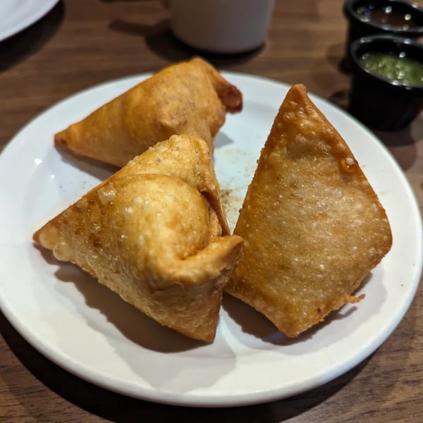 Three golden-brown Indian samosas on a white plate, with two small sauce bowls in the background.