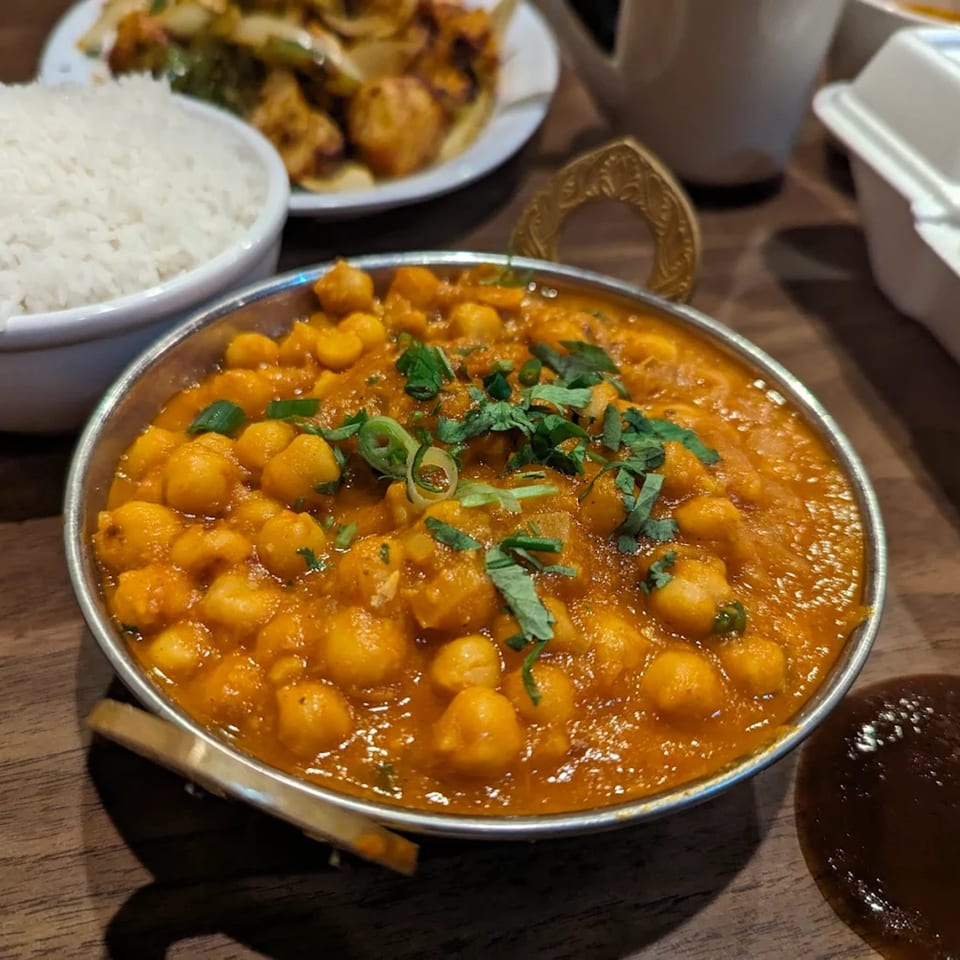 A metal bowl of chana masala, garnished with cilantro, next to white rice and other Indian dishes.