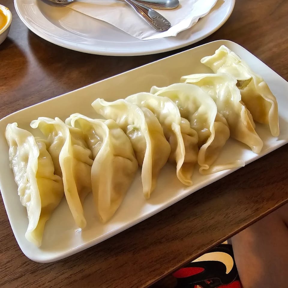 A white rectangular plate filled with steamed dumplings, with a white plate and cutlery in the background.